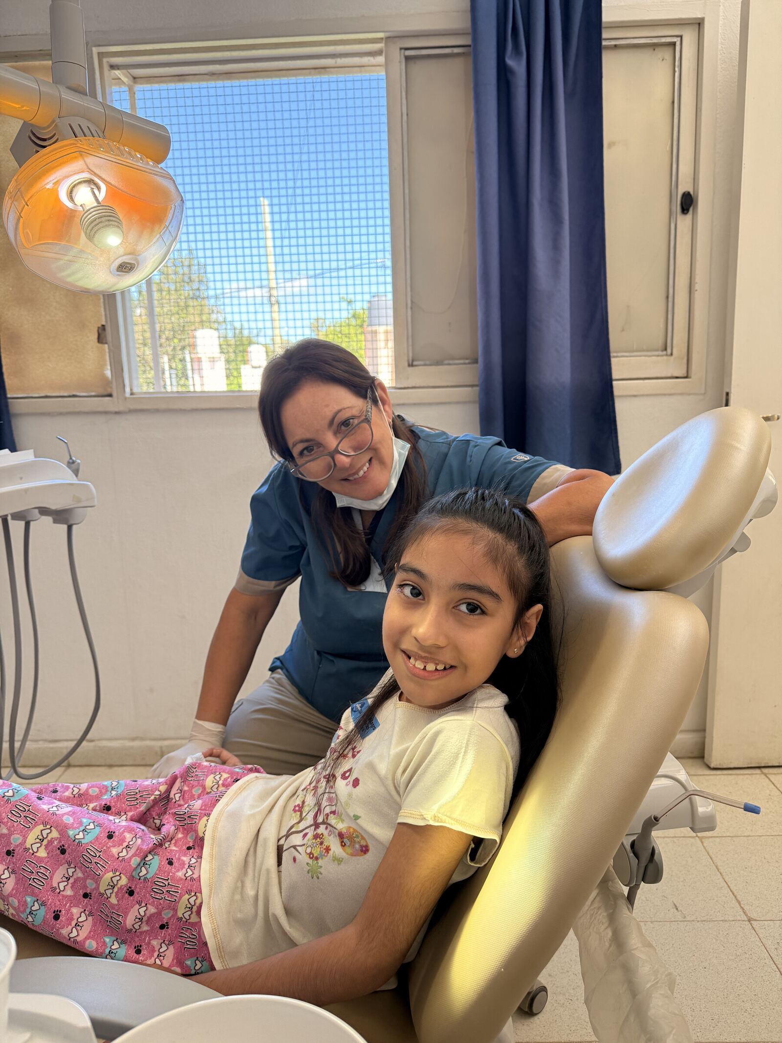 Una nena en el sillón odontológico sonríe a cámara con la odontóloga detrás.