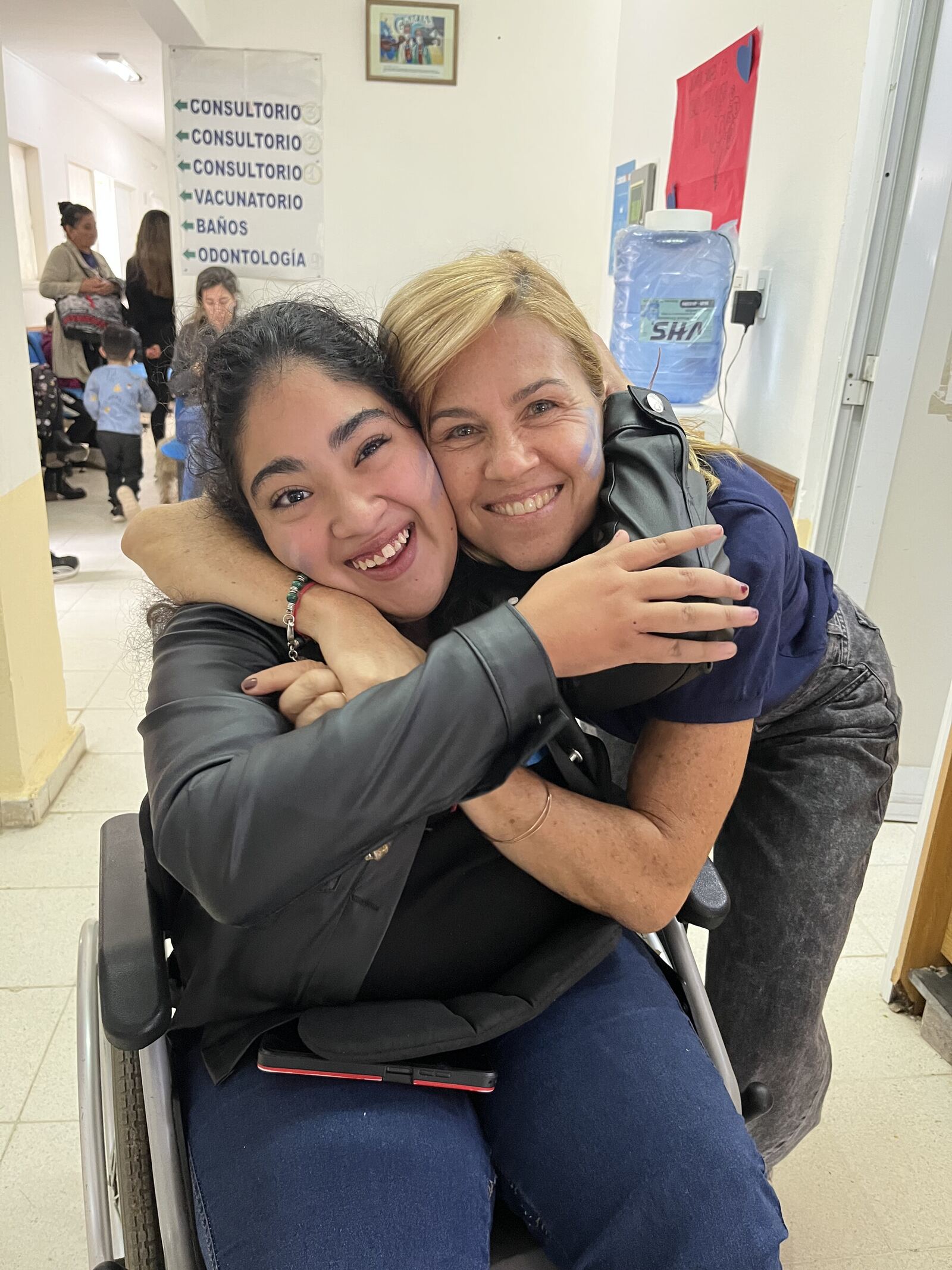 Una adolescente en silla de ruedas abraza a una profesional en el pasillo de los consultorios.