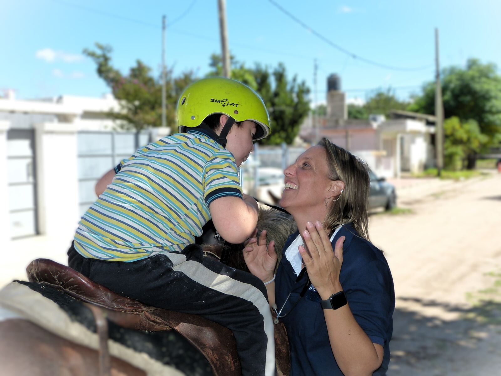 Equinoterapia en la calle del paraje: el nene con casco sobre el caballo y la terapeuta riéndose con él.