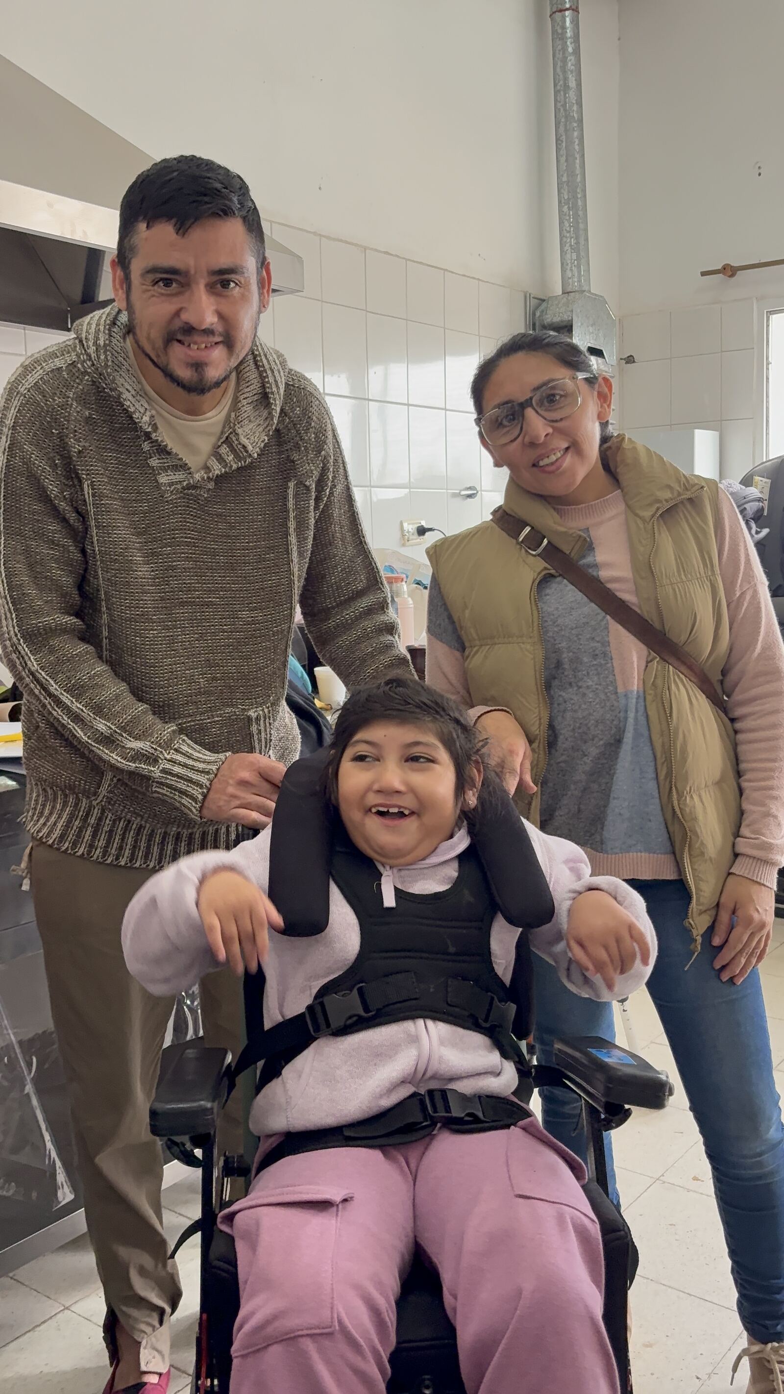 Un papá y una mamá detrás de su hija en la silla postural nueva; la nena se ríe a cámara